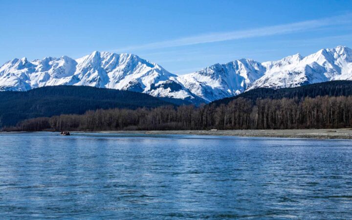 A raft of Alaska travelers floating down the Chilkat River while on a small ship cruise in Alaska with blue skies and the Chilkat mountains in the background.