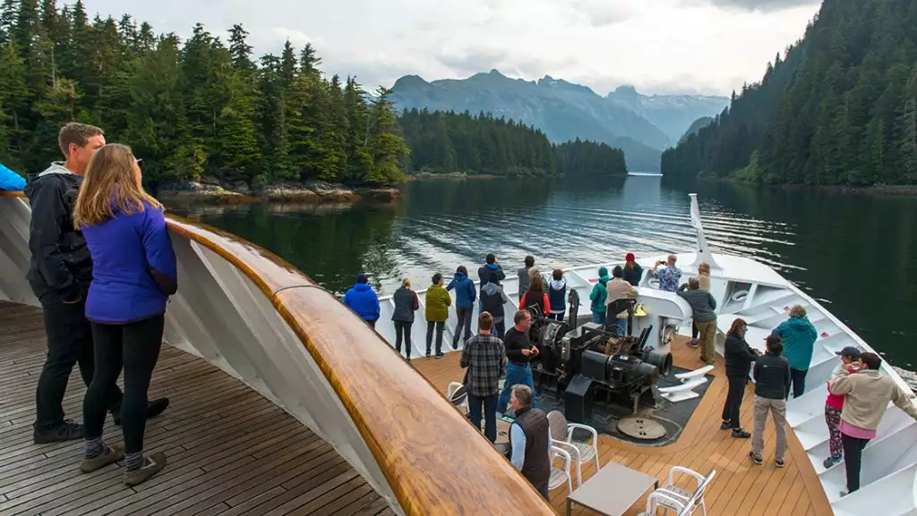Guests stand on decks of small ship while sailing calm inlet on the Glacier Bay National Park with Inside Passage & San Juan Islands cruise.