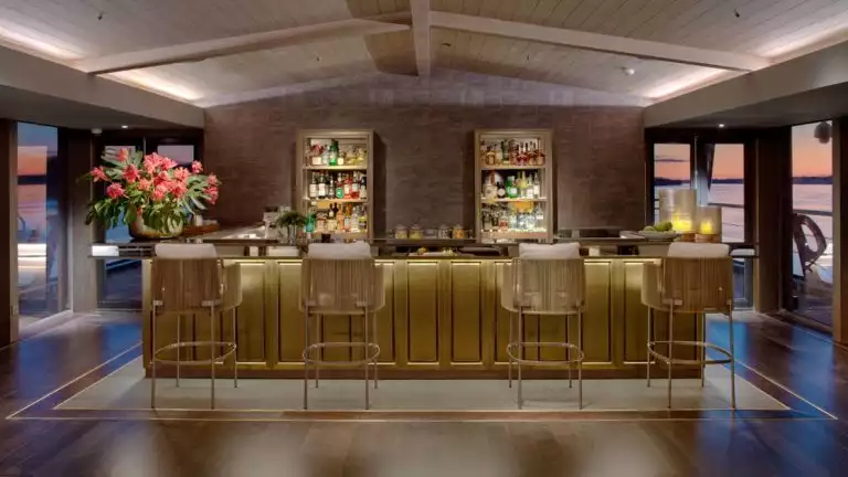 An illuminated gold bar with 4 tall bar chairs, two wall shelfs with alcohol bottles, surrounded by floor to ceiling windows.