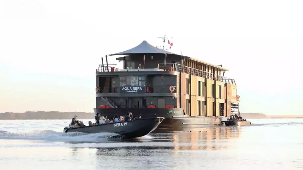 In the Peruvian Amazon, the modern three story Aqua Nera ship floats in a river as smaller metal passenger boats cruise around it.