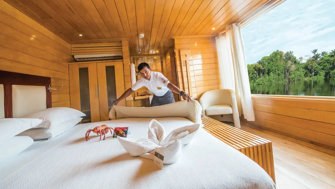 Cabin steward makes up king bed by large picture window in Guest Suite of Delfin II riverboat on an Amazon River cruise.