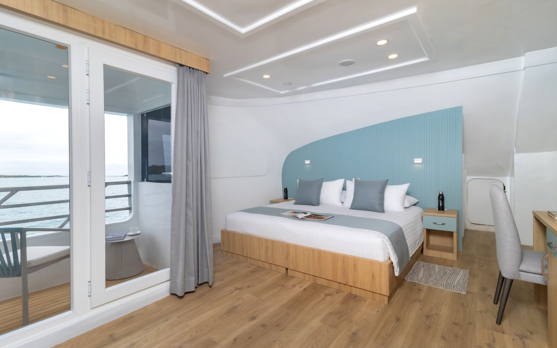 Main Deck Suite aboard Galapagos Sea Star room with double bed, balcony, and desk area. Tans, greys and blues for design