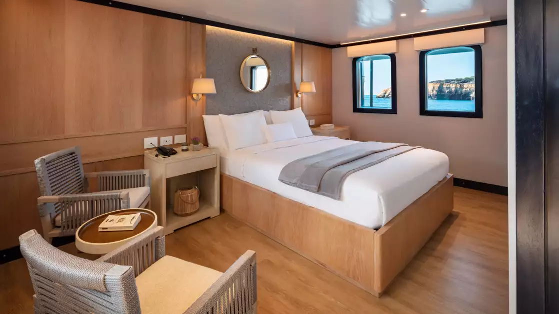 Large master suite with white bed linens surrounded by light wood finishes flanked by two windows and seating area