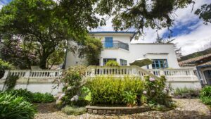 A guesthouse inside Hacienda Cusin, a 17th century estate found in Ecuador's Andes. White walls with bright blue window treatments and roof match the blue clounded sky, framed by lush green garden and foliage.