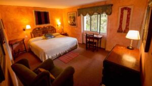 Warm glow of three lamps light this guestroom at Hacienda Cusin, a window opens to the garden, a small wooden table and chair, and a comfy lounge chair. Stay here as part of an Ecuador land tour.