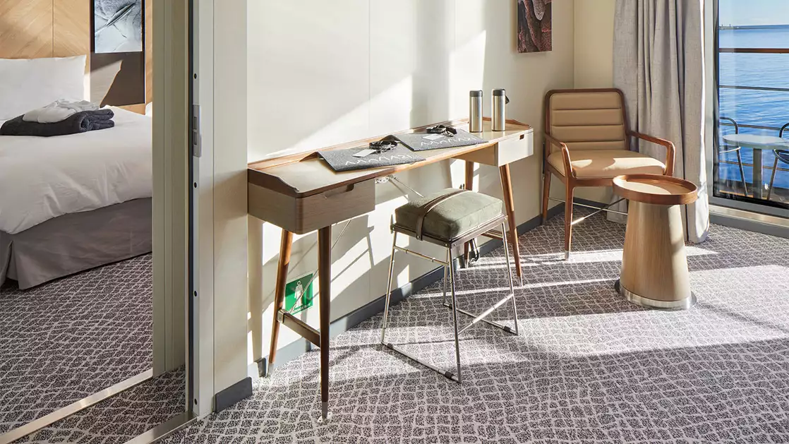 Corner of shared area with desk, cushioned stool & chair with side table overlooking balcony from Grande Suite on Magellan Explorer