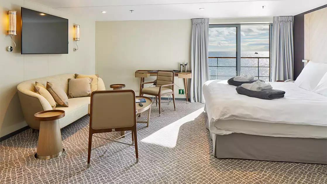 Penthouse Suite on Magellan Explorer with king bed, seating area with couch, 2 chairs, & desk beside sliding door leadng onto balcony