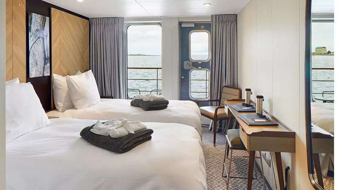 Veranda Cabin on Magellan Explorer with 2 twin beds across from desk with cushioned stool & chair in corner overlooking balcony