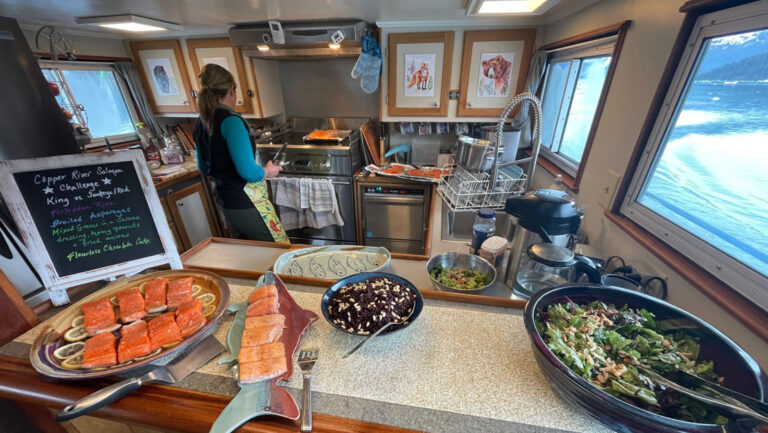Woman cooks fresh salmon in galley of M/V Discovery Prince William Sound yacht, with serving plate on bar beside green salad.