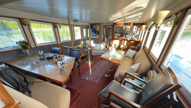 Lounge of M/V Discovery ship in Alaska, with 4-top tables, beige couches, window-lined walls, 3-stool bar & gray easy chair.