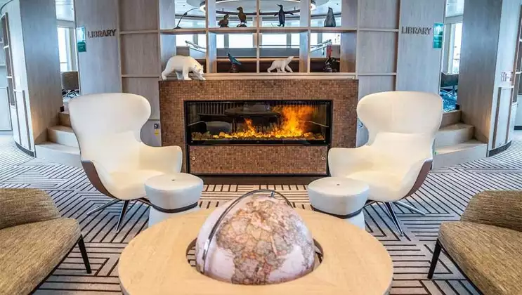 2 modern, white wingback chairs sit empty beside a coffee table with globe in front of a gas fireplace on National Geographic Endurance.