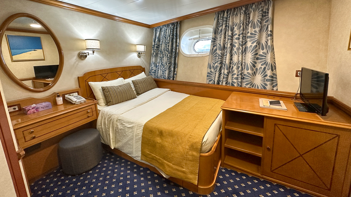Bed with towel shaped like a swan and small porthole, mirror and curtains aboard Panorama II