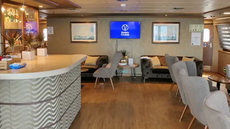 Bar of Panorama Mediterranean sailboat with silver & gold waves, wood floors, gray velvet chairs, TV & framed art.
