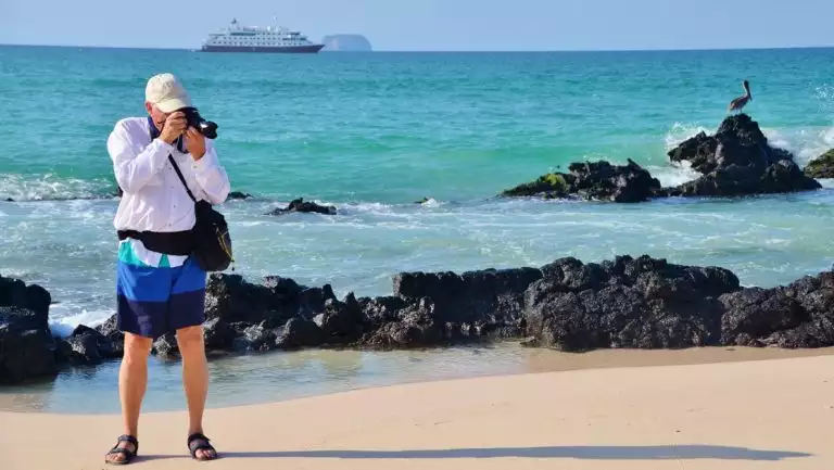 Man stands on a white-sand beach & photographs something out of frame with Santa Cruz II ship & pelican in background.