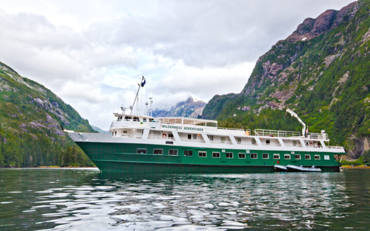 Portside exterior view of the Wilderness Adventurer small ship sitting in glassy water under a cloudy sky in Alaska.