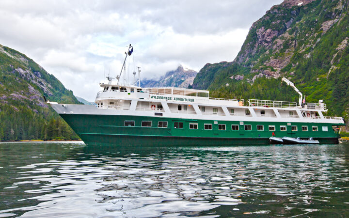 Portside exterior view of the Wilderness Adventurer small ship sitting in glassy water under a cloudy sky in Alaska.