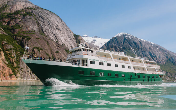 Wilderness Explorer small ship with white upper decks & green hull cruises in calm water on a sunny day.