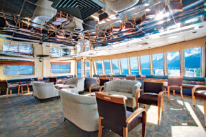 Lounge seating area with padded light blue loveseats & purple & wood chairs by windows aboard Wilderness Explorer
