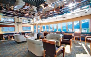 Lounge seating area with padded light blue loveseats & purple & wood chairs by windows aboard Wilderness Explorer