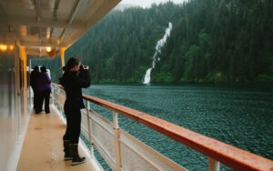 Woman in black pants & jacket looks through binoculars on side deck of Wilderness Explorer while cruising by a waterfall.