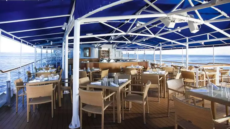 Al fresco dining area on La Pinta Galapagos yacht with rattan furniture, glass-top tables, blue cover & white structural poles.