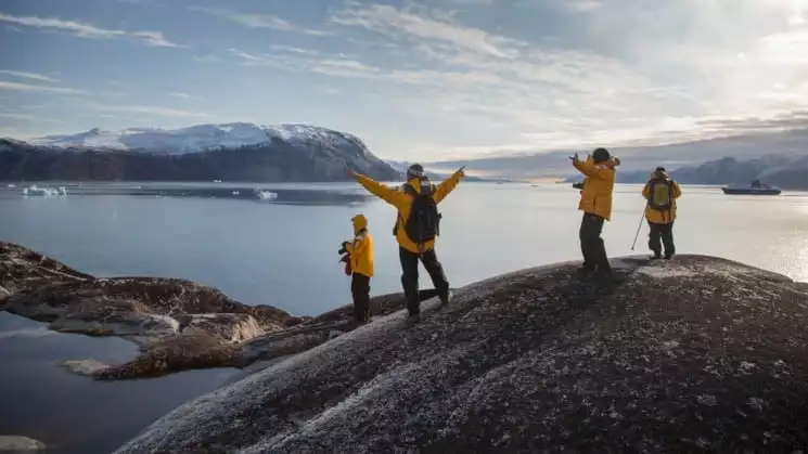 Guides and passengers from Essential Greenland expedition cruise stand atop a rock, arms outstretched, with a view of the ocean and the arctic in front of them.