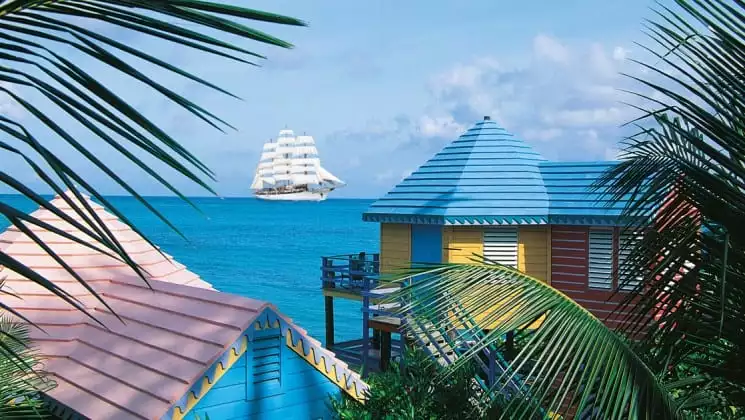 The small ship sea cloud sails on the tropical caribbean ocean with palm trees and yellow houses in the foreground