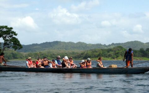 a group of adventure travelers rows a dugout canoe on flat water with mountains in the background on a sunny day during the best of panama land tour