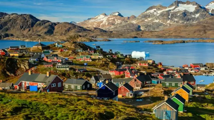 View of a village near the ocean in greeland with many red and green houses built around the rocky and hilly terrain.