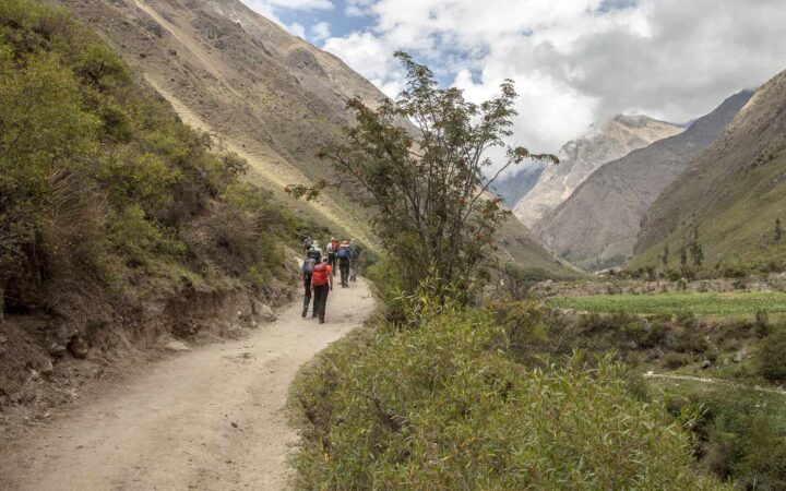 adventure travelers walk down a valley path on an overcast day with large mountains on either side during the inca trail trek
