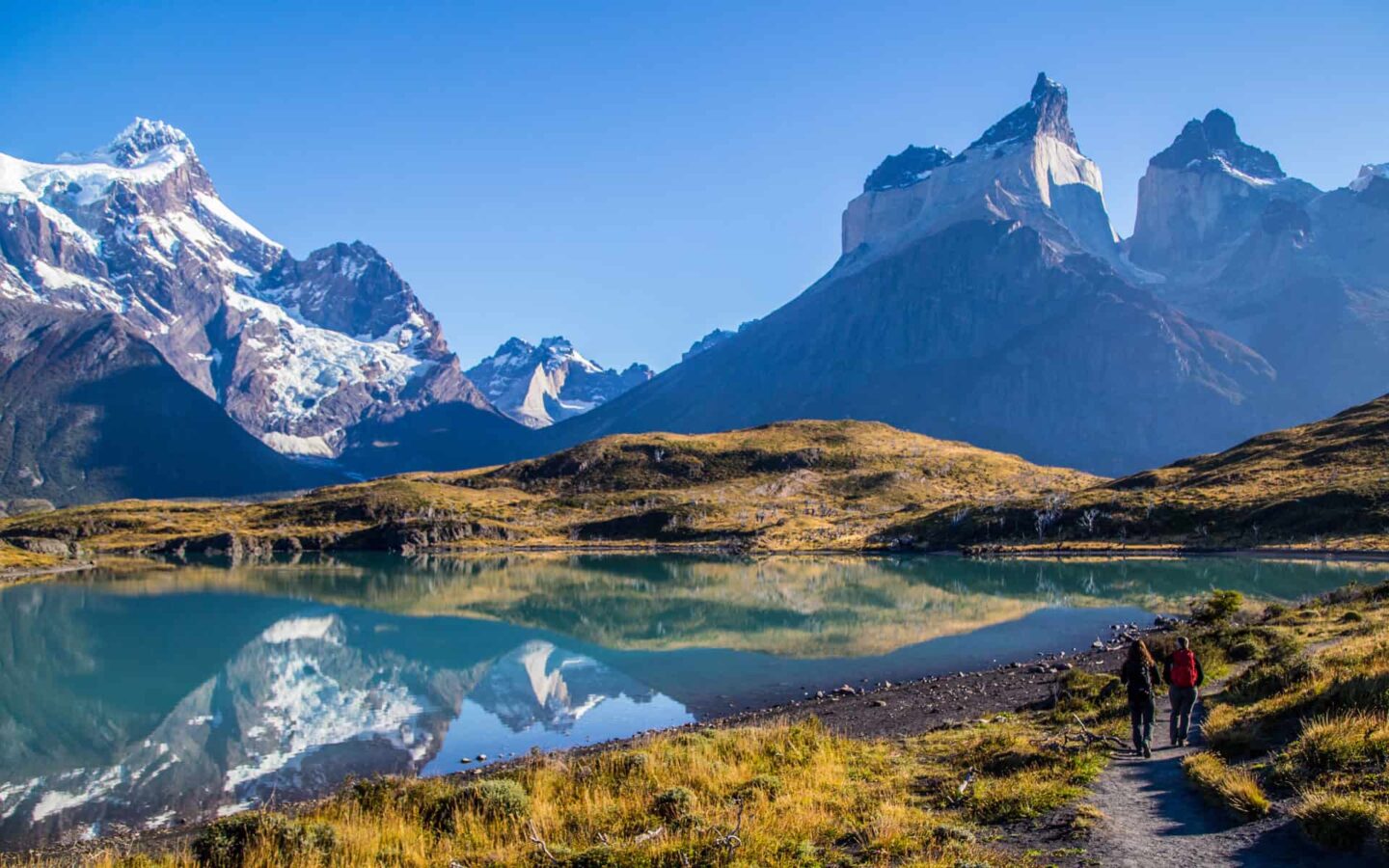 Gorgeous scene of a Patagonia adventure travel trip with a lake mirroring mountains and a trail on the shore.