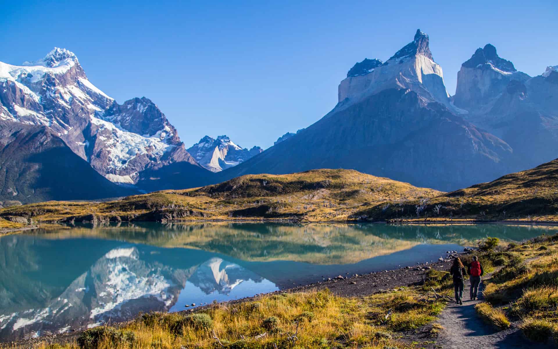 Gorgeous scene of a Patagonia adventure travel trip with a lake mirroring mountains and a trail on the shore.