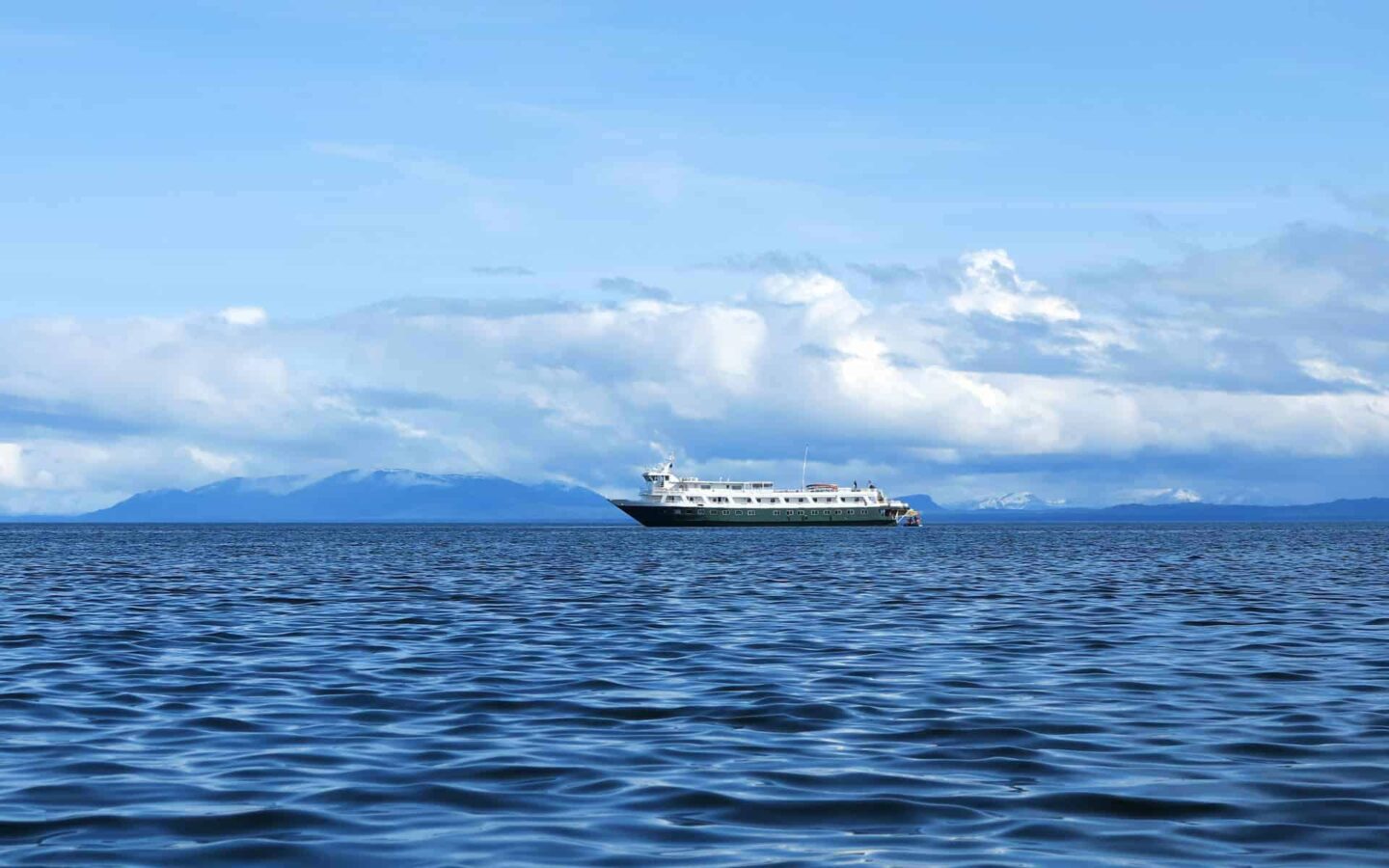 a lone alaska small ship cruises on calm water with clouds and mountains behind it on a mostly sunny day