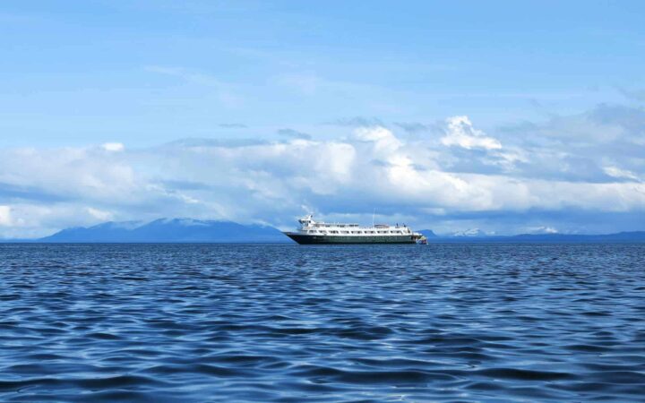 a lone alaska small ship cruises on calm water with clouds and mountains behind it on a mostly sunny day