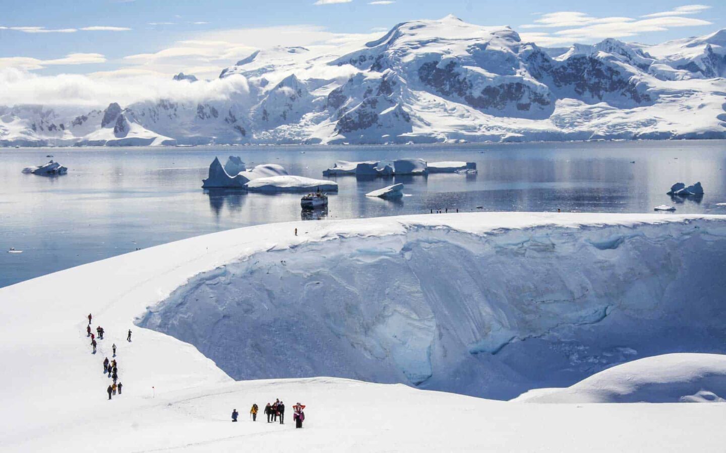 Travelers hike up a snowy ridge line with their small ship in the background.