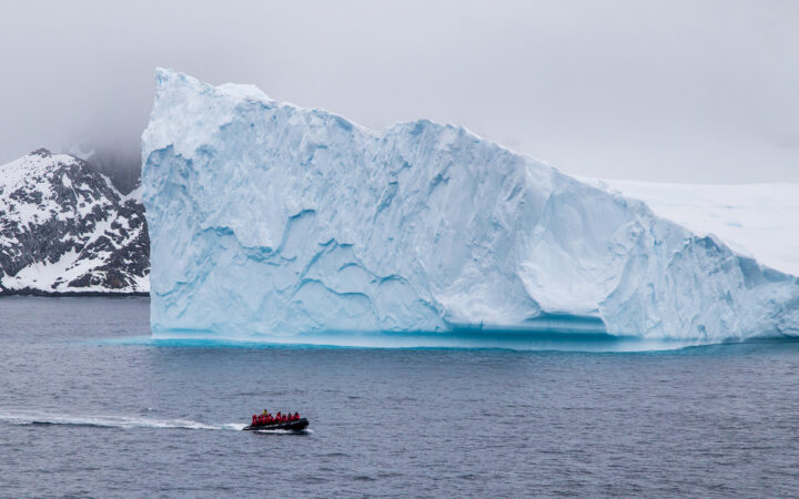 in Antarctica an inflatable skiff full of travelers in red parkas cruise by a massive floating iceberg the size of a building