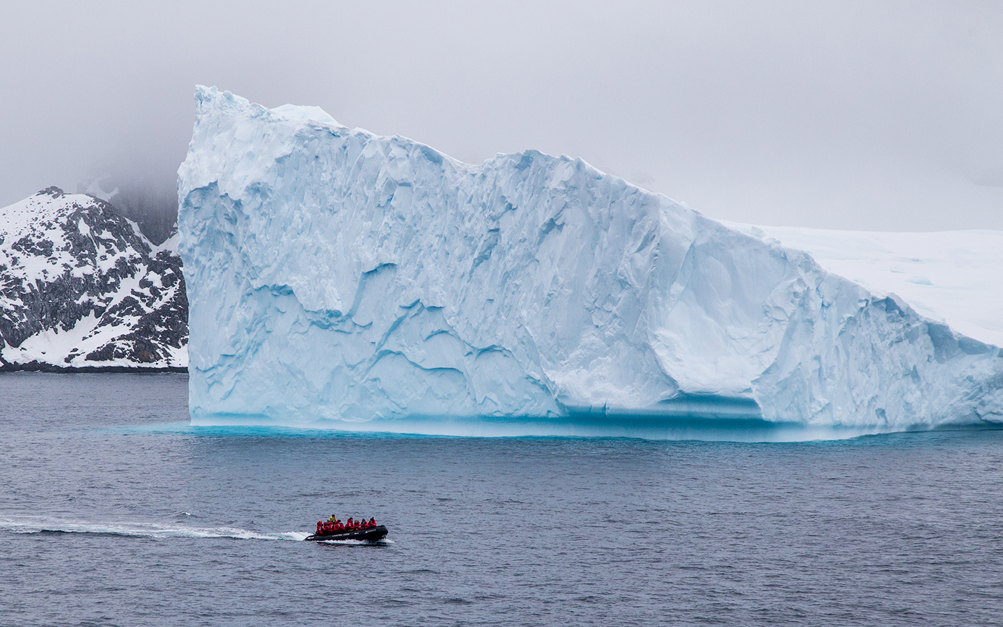 in Antarctica an inflatable skiff full of travelers in red parkas cruise by a massive floating iceberg the size of a building