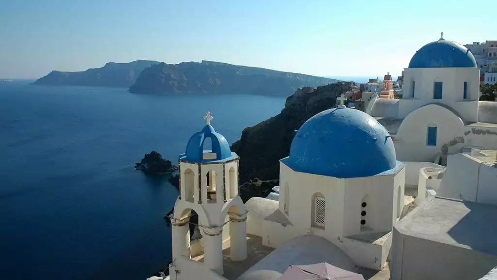 The blue-topped white buildings of Santorini Greece seen from above on a sunny day with the long mountainous coastline below them