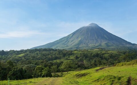 Arenal volcano from afar with bright green forest & grassland in the foreground, on a sunny day in Costa Rica.