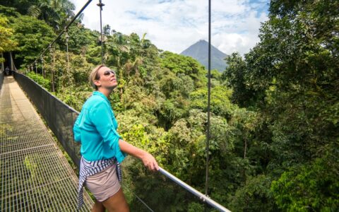 Woman in beige shorts & teal shirt stands on a hanging bridge in the Costa Rica rainforest & looks up into the trees on a sunny day.
