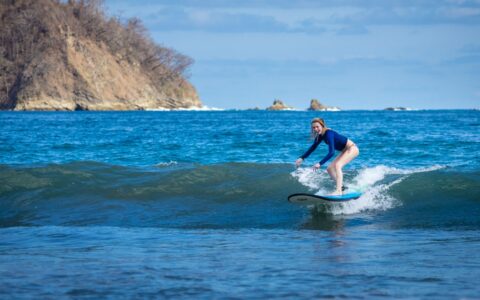 Girl surfs a small wave in bright blue ocean with a tan cliff behind on a sunny day during the Costa Rica Family Adventure.