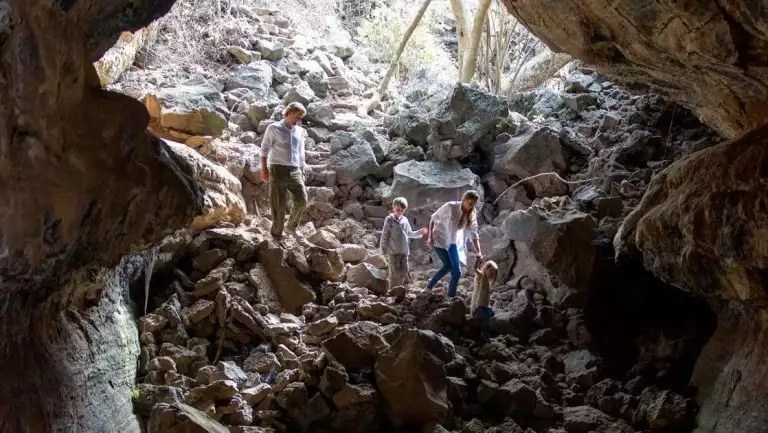 Man, woman & 2 kids walk down into a cave over large beige rocks under a sunny sky during a Galapagos Safari land tour.