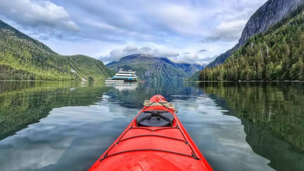 Tip of red kayak points to a small ship flaked by foresed green hillsides in Alaska.