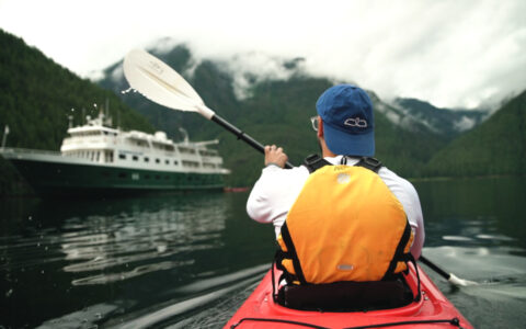Man paddles a red kayak toward a small ship with dark green hull & white upper decks backed by forested hills.