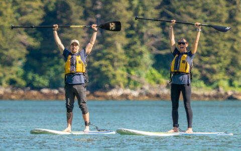 2 paddleboarders raise their paddles above their heads on a sunny day on the Glacier Bay National Park, Haines & Pelican cruise.
