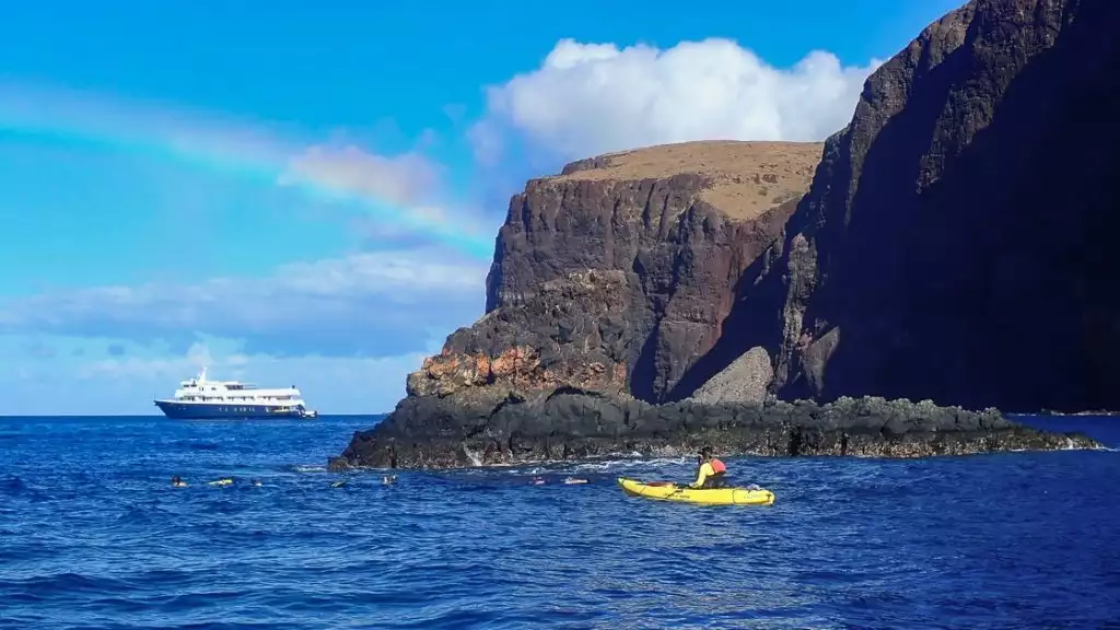 A rainbow across blue sky over a small ship on the horizon a single kayaker floats in the ocean near tall volcanic shoreline.
