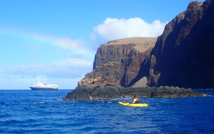 A rainbow across blue sky over a small ship on the horizon a single kayaker floats in the ocean near tall volcanic shoreline.