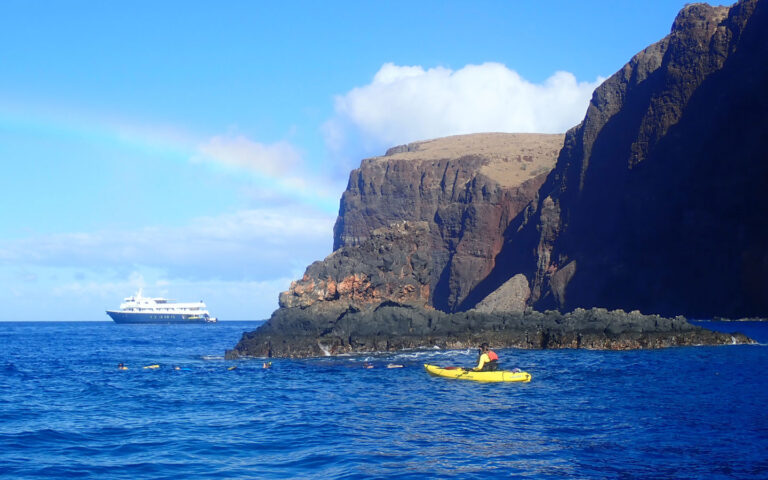 A rainbow across blue sky over a small ship on the horizon a single kayaker floats in the ocean near tall volcanic shoreline.