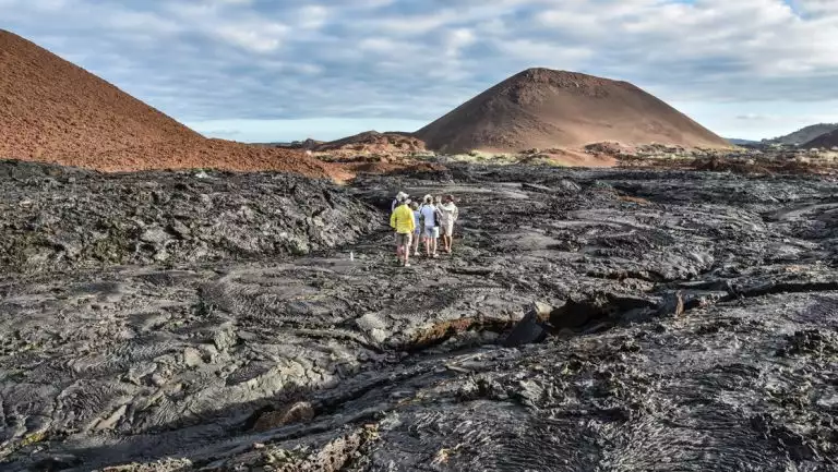 La Pinta Galapagos Cruise travelers walk over hardened black lava field near brown mountains on a cloudy day.