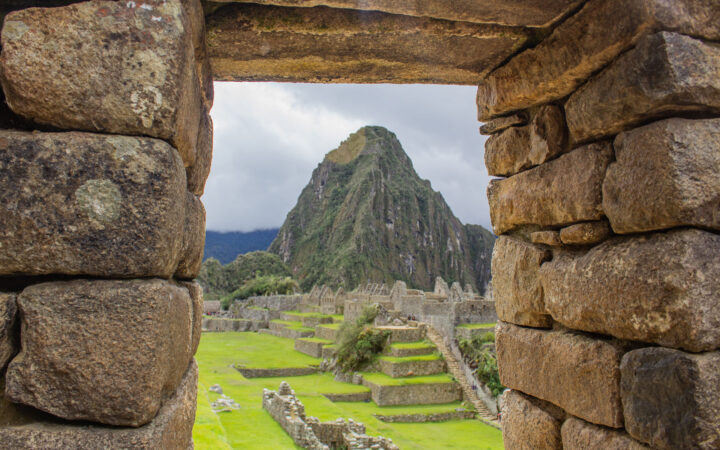 View through opening in brown stone wall at Machu Picchu citadel, looking onto bright green grassy levels & tall mountain behind.